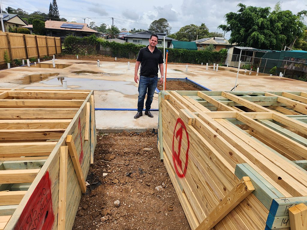 Tom Johnson standing on the slab at one of his rooming house builds, with timber wall frames stacked beside him
